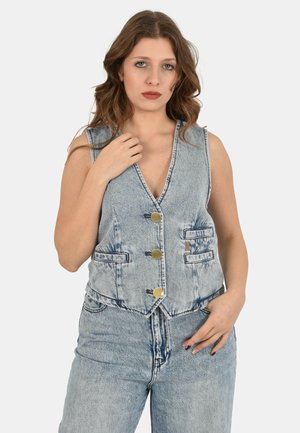 Donna con capelli mossi che indossa un gilet in denim azzurro chiaro senza maniche con bottoni dorati e jeans in denim a vita alta abbinati, in posa su sfondo semplice.