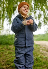 Morska vodootporna jakna i hlače s naglaskom u obliku zvijezde. Dijete nosi smeši pletenu kapu; ruke drže mali predmet vani.