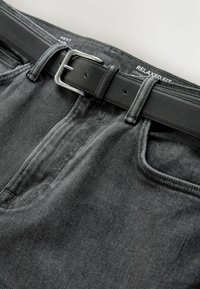Ceinture en cuir noir attachée à travers les passants sur un jean gris coupe décontractée, montrant la texture du tissu et les détails de la boucle métallique.