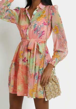 Frau trägt ein pastellfarbenes, geblümtes Kleid mit langen Ärmeln und einem Taillengürtel, dazu eine goldene Perlen-Schultertasche mit Kettenriemen.