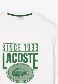 Camiseta de algodón blanca con un cuello redondo verde, texto en negrita verde que dice "DESDE 1933 LACOSTE" y un gráfico de cocodrilo verde.