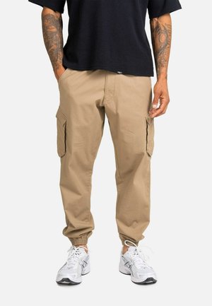 Mann trägt beige Cargo-Jogginghose mit elastischen Bündchen, weiße Sneakers und ein schwarzes kurzärmeliges Shirt, Hände in den Taschen.