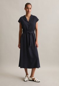 Marineblaue Midi-Kleid mit kurzen Ärmeln, Knopfleiste und verstellbarem Taillenzug. Das Model trägt weiße Schnürsandalen. Glattes, leichtes Material.