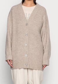 Cardigan beige de punto con cuello en V, textura acanalada, seis grandes botones y mangas anchas. Características de un ajuste oversize y material suave.