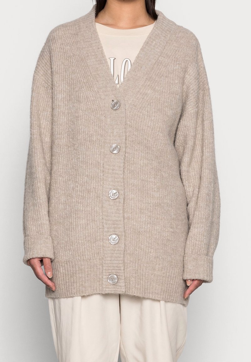 Cardigan beige de punto con cuello en V, textura acanalada, seis grandes botones y mangas anchas. Características de un ajuste oversize y material suave.