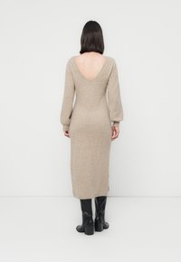 Robe beige à manches longues avec un dos nu, texture côtelée et fentes sur les côtés, associée à des bottes noires hauteur genou. Silhouette lisse et ajustée.