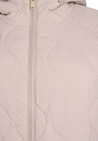 Lysebeige quiltet jakke med en glat tekstur, der har en central lynlås og en hætte. Designet inkluderer bølgede syningsmønstre.