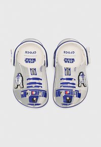 Baltos crocs su mėlynais akcentais ir R2-D2 dizainu, su ventiliacijos skylutėmis ir reguliuojama kulno juosta. „Star Wars“ logotipas viršuje.