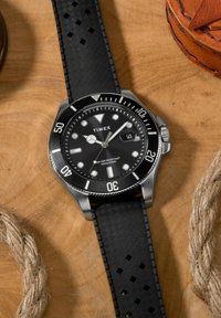 Timexi sukeldumiskell musta tekstuuritud silikoonrihmaga, musta sihverplaadi, roostevabast terasest korpuse, luminofoorliste markerite ja kuupäeva kuvaga.