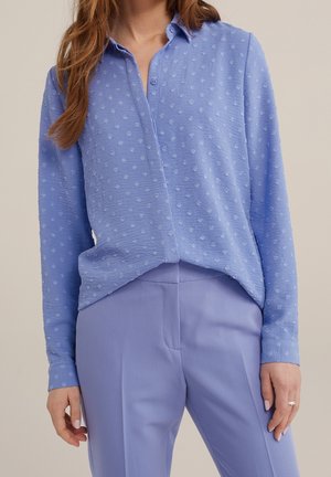 Vrouw draagt een lichtblauwe gestructureerde blouse met lange mouwen en kleine stippen, gecombineerd met een bijpassende lichtblauwe op maat gemaakte broek.