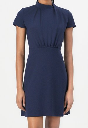 Granatowa sukienka z krótkim rękawem, wysokim dekoltem i marszczeniem w talii, noszona przez stojącą osobę na jednolitym tle.