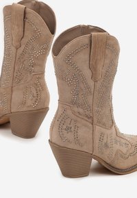 Bottes de cowboy en daim tan aux chevilles avec des clous en argent et des talons en bois empilés.