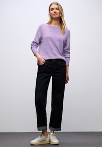 Lavenderfarbener Pullover mit entspannter Form und gerippten Bündchen, kombiniert mit dunkelblauen Jeans, die an den Bündchen hochgekrempelt sind, und hellen Sneakers.