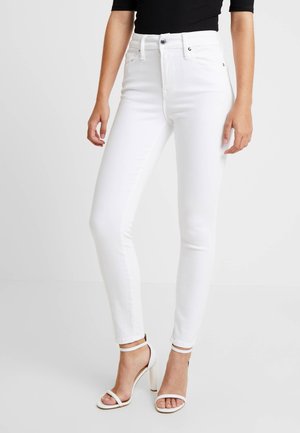 Donna che indossa jeans skinny bianchi a vita alta e sandali bianchi con tacco alto e punta aperta con cinturini alla caviglia, abbinati a un top nero.