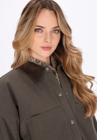 Giovane donna con lunghi capelli biondi mossi che indossa una giacca marrone con bottoni, colletto in velluto e una sciarpa a fantasia sotto.