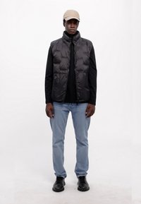 Μαύρο φορτισμένο γιλέκο με ψηλό γιακά, φορεμένο πάνω από ένα μαύρο μακρυμάνικο πουκάμισο, συνδυασμένο με ανοιχτόμπλε τζιν και μαύρα παπούτσια, επικεφαλής με ένα μπεζ καπέλο.