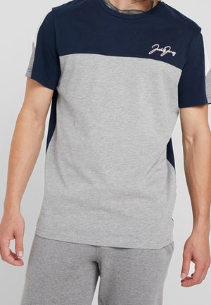 Mężczyzna w krótkim rękawie, koszulce w kolorze granatowym i szarym z logo „Jack & Jones” na piersi, połączonej z szarymi spodniami.