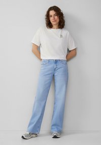 Balta, trumpomis rankovėmis marškinėliai su mažu logotipu kairėje krūtinės pusėje; derinama su šviesiai mėlynomis plačiomis kelnėmis ir pilkais sportiniais bateliais.