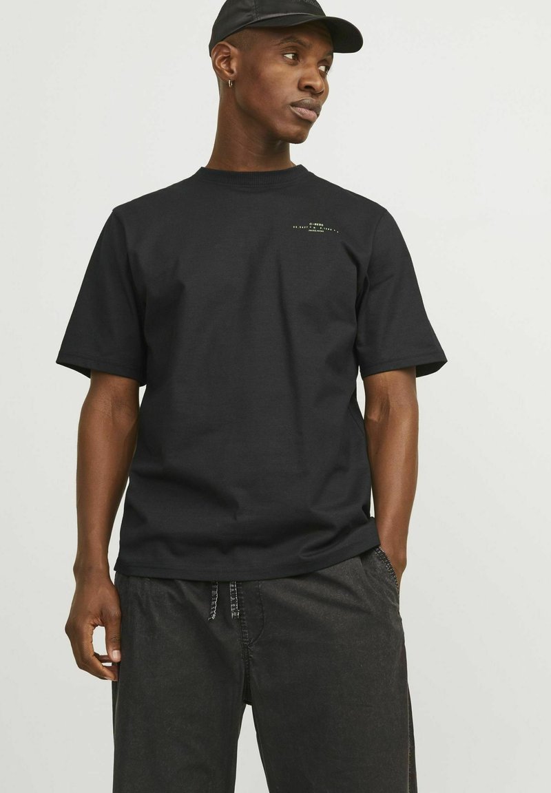 Black cotton t-shirt with short sleeves, crew neck, and subtle branding in contrasting color on the chest. Worn with dark pants.