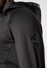 Melna sporta jaka ar gludu tekstūru, kurai ir apaļa logo uzpleču uz pleca un kapuce ar melnbaltu dekoratīvu apdari.