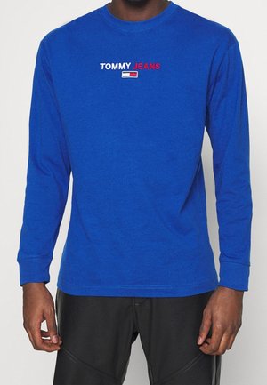 Homme portant une chemise à manches longues bleu royal avec le logo "TOMMY JEANS" et un emblème de drapeau sur la poitrine, assortie à un pantalon noir.