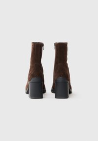 Bottines en suede marron avec un bout rond, un talon bloc noir discernable et un zip sur le côté. La vue arrière met en avant la hauteur et la forme.