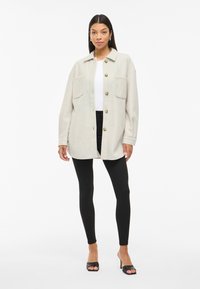 Ανοιχτό μπεζ overshirt ζακέτα με γιακά, δύο τσέπες στο στήθος και κλείσιμο με κουμπιά, συνδυασμένη με μαύρες κολάν και ανοιχτές γόβες.