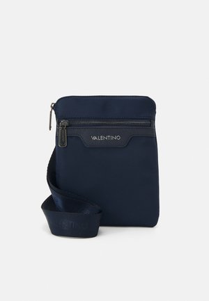 Sac bandoulière en nylon bleu marine avec une fermeture éclair sur le dessus, détail de logo à l'avant et une bandoulière texturée. Forme rectangulaire compacte avec surface lisse.