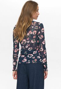 Donna con capelli lunghi che indossa una maglietta a maniche lunghe a fiori scuri e pantaloni larghi scuri, rivolta di lato con la maggior parte della schiena visibile.