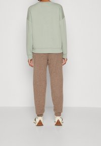 Sudadera verde claro, corte holgado, puños acanalados. Pantalones jogger marrones con textura, diseño cónico. Zapatillas deportivas de color neutro con suela de contraste.