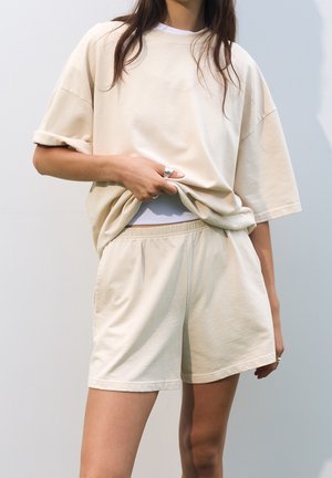 Femme portant un short beige ample et un T-shirt assorti oversize, soulevant légèrement le T-shirt avec une main.