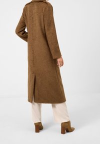 Brun, texturerad kappa med en lång silhuett och sidospalt, i kombination med ljusa byxor och bruna klackboots.