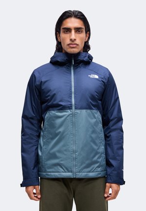 Homme portant une veste à capuche The North Face bicolore bleue, fermée par une fermeture éclair, debout devant un fond clair uni.