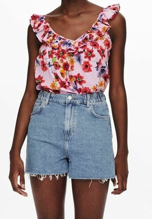 Femme portant un haut sans manches rose à fleurs avec volants et un short en jean bleu clair taille haute aux bords effilochés, debout sur fond blanc.