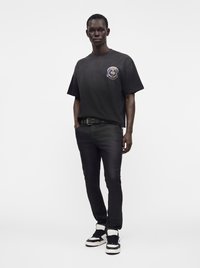 Άνδρας που στέκεται, φοράει μαύρο oversize T-shirt με γραφικό στο στήθος, μαύρα στενά τζιν, μαύρη ζώνη και μαύρα και άσπρα αθλητικά παπούτσια.