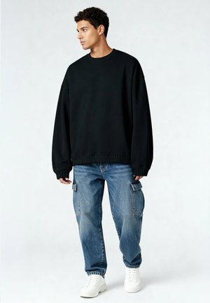 Felpa nera oversize con collo a giro, abbinata a jeans cargo azzurri chiari con tasche laterali e sneakers bianche. Look casual.