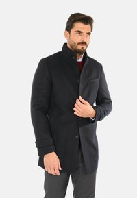 Cappotto di lana navy con colletto alto, dettagli con bottoni, una tasca petto e una vestibilità su misura. Tessuto liscio e design classico.