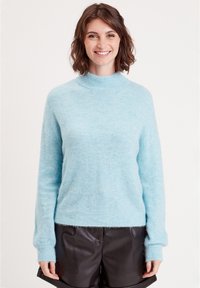 Pull bleu clair, duveteux avec un col haut et de longues manches, présentant des poignets et un ourlet côtelés. Associé à un short noir brillant.
