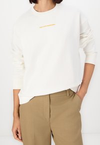 Sudadera de cuello redondo color gris claro con logo naranja "MarcoPoloDenim", corte relajado, puños acanalados, combinada con pantalones beiges. Material de algodón suave.