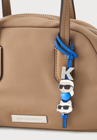 Brūna rokassoma ar sudraba "Karl Lagerfeld" plāksnīti un zilu ādas atslēgu piekariņu, kurā attēlots karikatūru Karl un kaķis, abi valkājot melnus saulesbrilles.