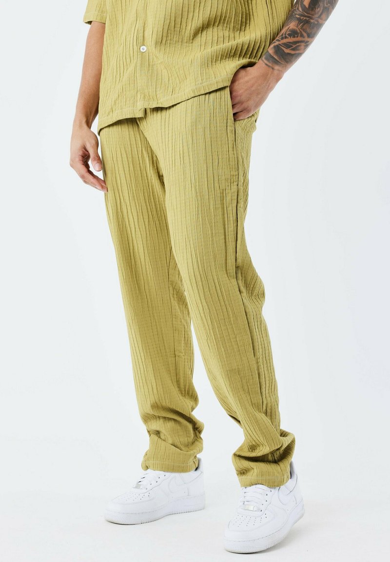 Pantalons texturés vert olive avec des rayures verticales, associés à des baskets blanches. Le tissu paraît léger et légèrement froissé.