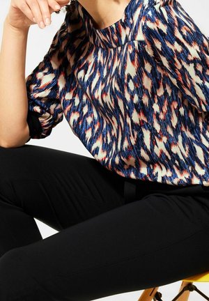 Blusa a fantasia con colori blu, rosso e crema, caratterizzata da una vestibilità ampia, maniche arricciate e scollatura rotonda. Abbinata a pantaloni neri.