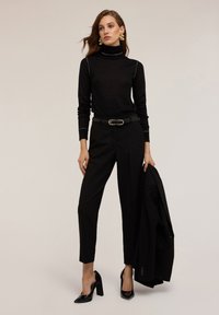 Pulover negru cu gât înalt și cusături contrastante, pantaloni negri eleganți, tocuri înalte negre elegante și un sacou negru aruncat pe un braț.