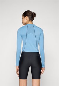 Lyseblå langermet topp med et teksturert bakpanel, kombinert med svarte tettsittende shorts. Materialet ser ut til å være elastisk og glatt.