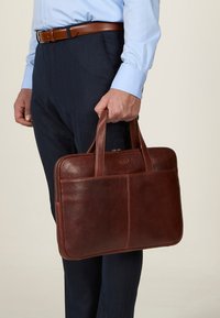 Valigetta in pelle marrone con due manici, design elegante, texture liscia, tasca frontale e chiusura con zip, tenuta da una persona in pantaloni eleganti.