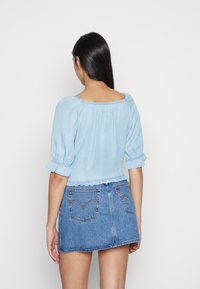 Blusa de ganga azul-clara, curta, com mangas bufantes e bainha franzida, combinada com uma saia mini de ganga com bolsos traseiros e logótipo distintivo.