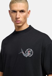 Sort bomulds t-shirt med et farverigt broderet koi-fisk design på venstre brystområde. T-shirten har en afslappet pasform og høj hals.