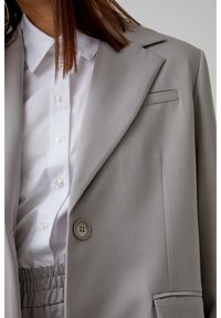 Grau maßgeschneiderte Blazer mit Reverskragen, einem einzelnen Knopfverschluss und einem subtilen strukturierten Stoff. Kombiniert mit einem weißen Hemd darunter.