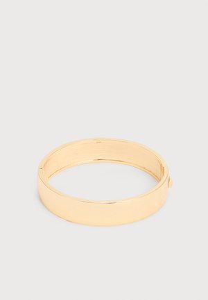 Βραχιόλι-μανσέτα από χρυσό μέταλλο με λεία, γυαλιστερή επιφάνεια και μικρό κούμπωμα σε απλό φωτεινό φόντο.