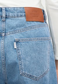 Ανοιχτό μπλε τζιν με πίσω τσέπη και καφέ δερμάτινη ετικέτα με την επιγραφή «Mads Nørgaard Copenhagen», φορεμένο με λευκό τοπ.